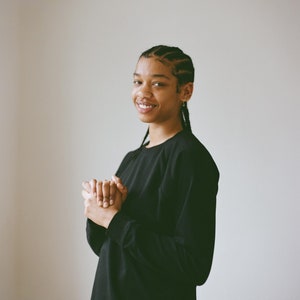 May include: A person wearing a black long-sleeve shirt smiles at the camera. The person has braided hair and is wearing a nose ring and earrings. The person is holding their hands together.