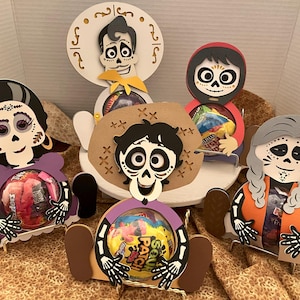 May include: Five colorful, cartoon-style, wooden candy holders featuring characters from the movie Coco. Each holder has a different character, including Miguel, Hector, Mama Imelda, Dante, and a skeleton with long braids. The holders are designed to hold candy or small treats.