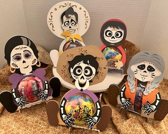 May include: Five colorful, cartoon-style, wooden candy holders featuring characters from the movie Coco. Each holder has a different character, including Miguel, Hector, Mama Imelda, Dante, and a skeleton with long braids. The holders are designed to hold candy or small treats.
