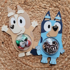 May include: Two handmade paper dog-shaped candy holders, one tan and one blue, each with a clear bubble containing wrapped candies. The tan dog has a white face and brown accents, while the blue dog has a white face and black accents. Both are on a woven surface.