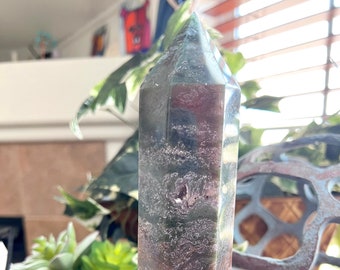 May include: A green, brown, and white agate crystal point with a faceted shape. The crystal is on a black circular stand.