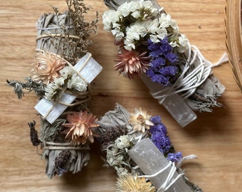 May include: Three white sage smudge sticks decorated with dried flowers and clear quartz crystals. The smudge sticks are wrapped with twine and adorned with dried flowers in shades of white, purple, and pink.