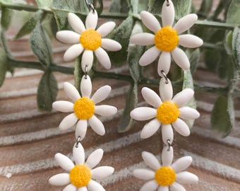 May include: Daisy flower earrings with white petals and yellow centers. The earrings are made of a lightweight material and hang from silver-colored hooks. The earrings are a dangle style and are a floral theme.