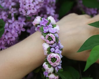 May include: A purple and white flower bracelet made of polymer clay. The bracelet is made up of small flowers and leaves, and is worn on a person's wrist.