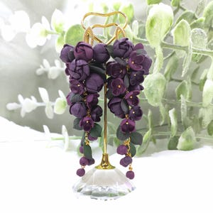 Puede incluir: Pendientes colgantes con racimos de flores moradas y hojas verdes. Los pendientes cuelgan de aros y cadenas doradas. Se exhiben en un soporte dorado con una base transparente facetada.