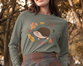 May include: Sage green long-sleeve sweatshirt featuring a cartoon hedgehog surrounded by autumn leaves, berries, and an acorn. The model is wearing a brown and red plaid skirt. The background is a blurred view of trees.