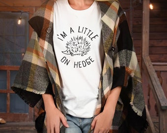 Könnte beinhalten: Ein weißes T-Shirt mit einer schwarzen Grafik eines Igels und dem Text "I'm a little on hedge". Die Person, die das Shirt trägt, trägt auch einen karierten Schal.