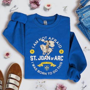May include: A royal blue sweatshirt with a white graphic design featuring a woman on horseback. The graphic includes the text "I am not afraid", "St. Joan of Arc", "I was born to do this", and the numbers "14" and "31".