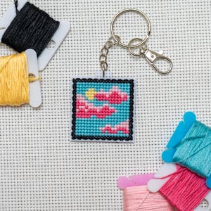 Op de afbeelding: Een vierkante kruissteek sleutelhanger met een luchtscène met roze wolken en een gele zon tegen een blauwe achtergrond. De sleutelhanger heeft een zwarte rand en een zilveren ring. Spoelen met kleurrijk borduurgaren liggen rond de sleutelhanger.