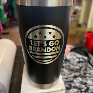 May include: Black stainless steel tumbler with a silver 'Let's Go Brandon' design. The design includes five stars and a red, white and blue stripe.