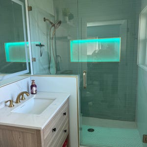 May include: A contemporary bathroom featuring a glass shower enclosure, a wooden vanity with a white countertop, and gold fixtures. The shower has teal-coloured tiles and a built-in shelf with a green light. The vanity has a white sink and a gold tap.