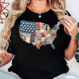 May include: Black t-shirt featuring a patchwork design of the United States map. The map incorporates the American flag with blue and white stars, and colorful patches representing different states. The shirt is a casual style.