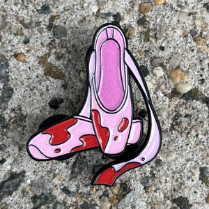 May include: A pink and black enamel pin shaped like a ballet slipper with red blood-like accents. The pin is set against a textured gray background.