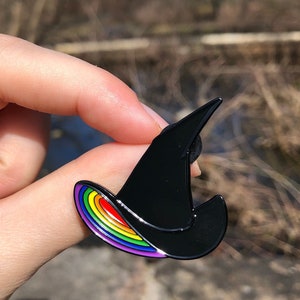 May include: A black witch hat enamel pin with a rainbow stripe design.