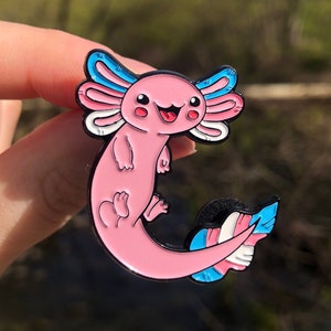 Op de afbeelding: Een roze axolotl-emaille pin met blauwe en witte accenten. De axolotl glimlacht en heeft een trans pride-vlagstaart.