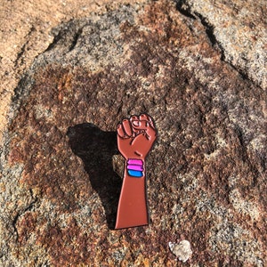 Op de afbeelding: Een bruine emaille pin met een gebalde vuist met een roze, blauw en paars gestreept ontwerp op de arm. De pin is geplaatst op een getextureerd, bruin en tan rots oppervlak.