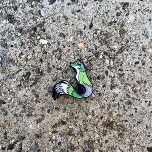 Könnte beinhalten: Ein farbenfroher Emaille-Pin in Form eines Fuchses. Der Fuchs hat einen grünen Kopf und Körper, einen weißen und grauen Schwanz und eine schwarze Umrandung. Der Pin befindet sich vor einem strukturierten, grauen Hintergrund.