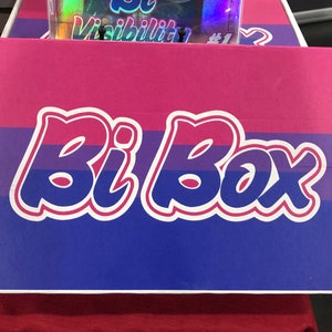 May include: A pink, blue and purple box with the text "Bi Box" in white lettering. The box has a bisexual pride flag design.