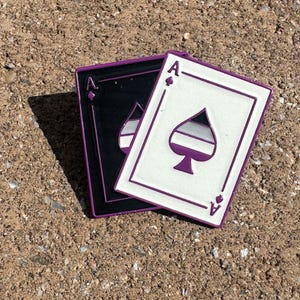 Peut inclure: Deux broderies en émail rectangulaires en forme de cartes à jouer. L'une est noire avec des contours violets et un symbole de pique violet. L'autre est blanche avec des contours violets et un pique rempli de bandes horizontales noir, gris, blanc.