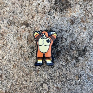 Op de afbeelding: Een kleurrijke emaille pin met een cartoon beer. De beer is afgebeeld met oranje, gele en bruine accenten, met een zwarte pootafdruk op de borst. De pin heeft een zwarte rand en is geplaatst tegen een getextureerde, grijze achtergrond.