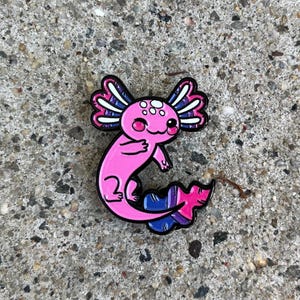 Könnte beinhalten: Ein pinkfarbener Axolotl-Emaille-Pin mit schwarzen Umrissen. Der Axolotl hat weiße, rosa und blaue Akzente an seinen Kiemen und seinem Schwanz, mit einem Bi-Pride-Flaggen-Design auf dem Schwanz. Der Pin befindet sich auf einer strukturierten, grauen Oberfläche.