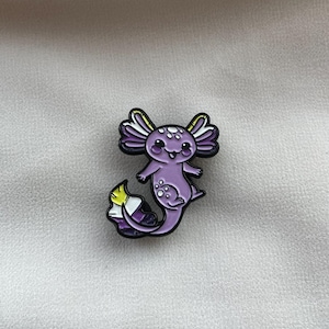 May include: A purple axolotl enamel pin with black outlines and a non-binary flag design. The axolotl has a smiling face, white spots, and purple gills. The pin is on a white, textured surface.
