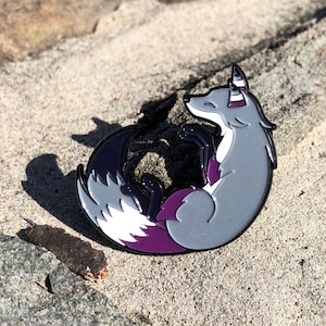 May include: A gray fox enamel pin with white and purple accents, curled up in a relaxed pose. The pin has a black outline and is set against a sandy background. The fox's tail and ears feature the purple and white colors.