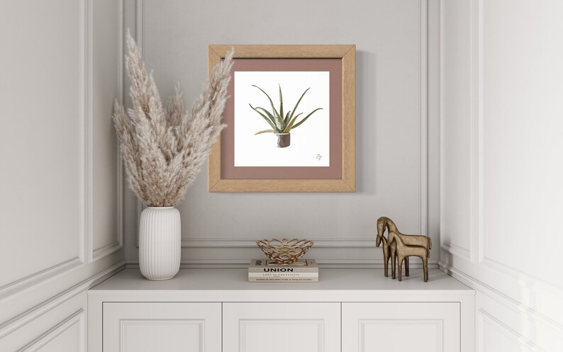 Puede incluir: Una impresi&oacute;n enmarcada de una planta de aloe vera verde en una maceta marr&oacute;n sobre un fondo blanco. El marco es de madera marr&oacute;n claro con un paspart&uacute; rosa claro. La impresi&oacute;n est&aacute; en un estante blanco con un jarr&oacute;n blanco con hierba de la pampa seca, un cuenco de metal dorado y dos figuritas de caballos de metal dorado.