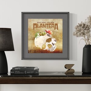 Op de afbeelding: Een ingelijst drukwerk met een schedel en bloemen op een textuur achtergrond. De tekst "REAP WHAT YOU SOW PLANTERA" is in bruin op de print gedrukt.