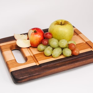 May include: A rectangular wooden cutting board with a handle, featuring alternating stripes of light and dark wood. Apples and grapes are arranged on the board. The board has a dark brown border and a groove for catching juices. The fruit includes a red apple, a green apple, and red and green grapes.