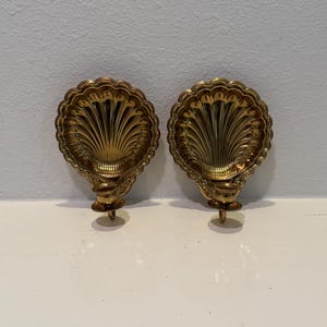 Könnte beinhalten: Zwei verzierte, goldfarbene Wandleuchter. Jeder Leuchter hat ein muschelförmiges Design mit detaillierten Rippen und einem kleinen, dekorativen Sockel. Die Leuchter sind auf einer weißen Oberfläche montiert.