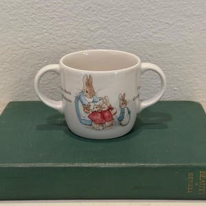 Puede incluir: Una taza de cerámica blanca con dos asas que presenta una ilustración colorida de Peter Rabbit y otros conejos con ropa roja y azul. La taza tiene texto impreso en el lateral y está diseñada con dos asas simétricas en lados opuestos del recipiente.