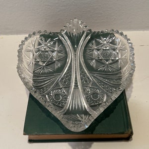 May include: A clear glass heart-shaped dish with a cut glass design. The dish has a raised handle in the center.