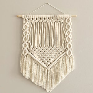 Puede incluir: Un tapiz de macramé blanco con un patrón geométrico y flecos. El tapiz está sujeto a una varilla de madera.
