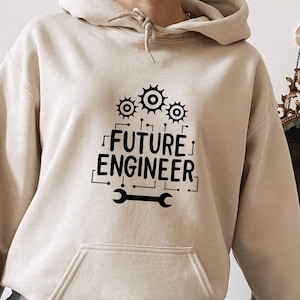 May include: A beige hoodie with a black graphic print that says "FUTURE ENGINEER" with gears and a wrench. The hoodie is worn by a person sitting on a white stool.