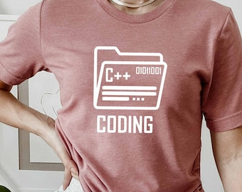 Puede incluir: Una camiseta rosa con un gráfico blanco de una carpeta etiquetada como "C++" y "Coding".