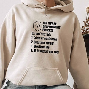 Könnte beinhalten: Ein beiger Hoodie mit einem schwarzen Grafikdruck, der "SOFTWARE DEVELOPMENT PROCESS" lautet und die Schritte auflistet: "0. I can't fix this", "1. Crisis of confidence", "2. Questions career", "3. Questions life", "4. Oh it was a typo, cool".