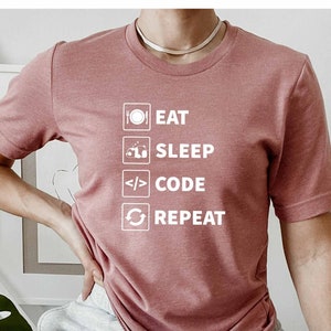 Könnte beinhalten: Ein pinkfarbenes T-Shirt mit einer weißen Grafik, auf der "Essen, Schlafen, Programmieren, Wiederholen" steht. Die Grafik enthält Symbole für einen Teller, ein Bett, Code und ein Aktualisierungssymbol.