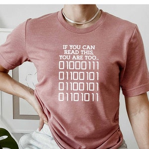 Könnte beinhalten: Ein pinkfarbenes T-Shirt mit dem Text "If you can read this, you are too." und einer Reihe von Binärcode: "01000111 01100101 01100101 01101011".