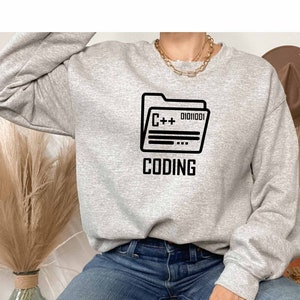Puede incluir: Una sudadera gris con un gráfico negro de una carpeta con el texto "C++ 01010001 CODING" en ella.