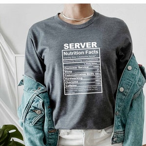 Puede incluir: Una camiseta de manga larga gris oscuro con un gráfico blanco que dice "SERVER Nutrition Facts" y enumera varios valores "nutricionales" para un servidor, como el servicio al cliente, el sueño, las habilidades de comunicación, el trabajo duro, el carisma y la cafeína.