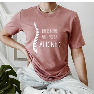 May include: A pink t-shirt with a white graphic of a spine and the text "Life is better when you're aligned".