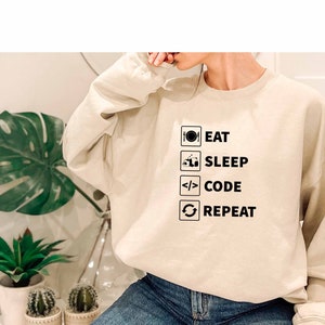 Könnte beinhalten: Ein beiger Pullover mit einer schwarzen Grafik, auf der "Essen, Schlafen, Programmieren, Wiederholen" steht.