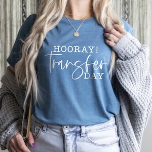 Puede incluir: Una camiseta azul con el texto "HOORAY! Transfer Day" en letras blancas.
