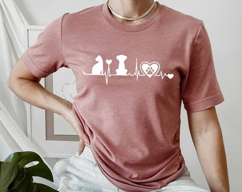 May include: A woman wearing a dusty rose t-shirt with a white graphic of a cat, dog, and heart with a paw print inside. The graphic is a heartbeat line with the animals and heart on the line.
