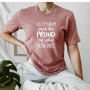 May include: Dusty rose-colored t-shirt with the text "KEEP CALM and be PROUD of who YOU ARE" in white. The shirt is short-sleeved and has a crew neck. The person is wearing gray sweatpants and white socks.