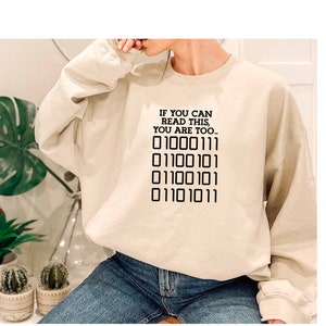 Könnte beinhalten: Ein beiger Pullover mit dem Text "Wenn du das lesen kannst, bist du auch..." und einer Reihe von Binärcode: "01000111 01100101 01100101 01101011 01101011".