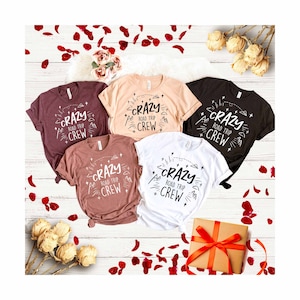 May include: Five t-shirts with the text "Crazy Road Trip Crew" in black lettering on a white background. The shirts are in various colors, including black, white, pink, and brown.