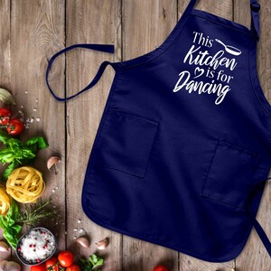 Puede incluir: Un delantal de cocina azul marino con texto blanco que dice "This Kitchen is for Dancing" y un pequeño icono de sartén blanco. El delantal tiene dos bolsillos.