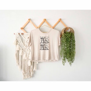 Può includere: Una maglietta beige con una grafica nera di un omino stilizzato davanti a un computer. Il testo sulla maglietta recita "the code doesn't work... why? the code works... why?"